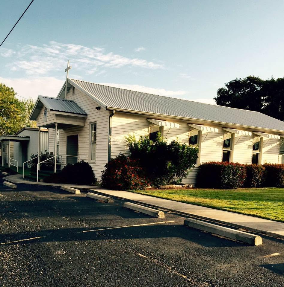 The Kingsbury Methodist Church sanctuary: a white wood-sided building with a standing-seam metal roof, a cross at the peak, and a covered entry porch with a ramp, seen from the parking lot in morning light.